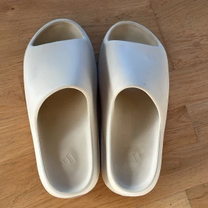 Adidas Yeezy Slide beige - Adidas Yeezy Slide i beige med chunky, räfflad sula och öppen tå. Slidesen har en minimalistisk design och är tillverkade i syntetmaterial som ger en mjuk och bekväm känsla. Perfekt för dig som gillar streetwear och vill ha något stilrent till sommaren.