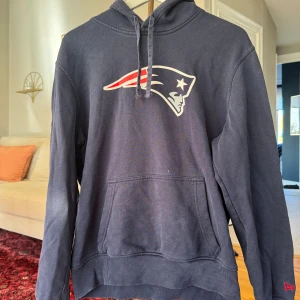 Mörkblå Patriots hoodie från New Era - Säljer en mörkblå hoodie från New Era med New England Patriots-logga på bröstet. Tröjan har huva med snörning, magficka och långa ärmar. Perfekt för dig som gillar amerikansk fotboll och vill ha en sportig look.