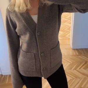 Brun kofta med fickor och knappar - Mysig brun kofta med djup v-ringning, stora knappar framtill och två ribbade fickor. Tröjan har långa ärmar och breda ribbade muddar vid ärmslut och nederkant. Perfekt att slänga på sig när det är kyligt ute.inte min bild men fråga så skickar jag mina egna bilder,den är inte använd 