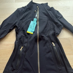 Svart sportig vindjacka med guldzip liknanade lulu - Svart, figursydd vindjacka från Zhen Yang med guldig dragkedja och detaljer. Jackan har långa ärmar, två fickor med dragkedja och hög krage. Materialet är lätt och stretchigt, perfekt för en aktiv stil. Snygg och clean look med sportiga vibbar. 