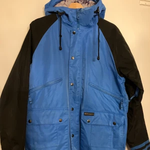 Blå och svart vindjacka från Burton - Snygg blå och svart vindjacka från Burton med huva och coola detaljer. Jackan har dragkedja och tryckknappar framtill, snörning i huvan och flera fickor med dragkedja. Fodret har ett unikt tryck och jackan är perfekt för dig som gillar streetwear.