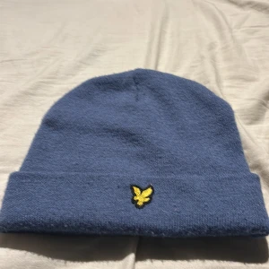 Blå mössa från Lyle & Scott Junior - Snygg blå mössa från Lyle & Scott Junior med klassisk gul broderad örn-logga framtill. Mössan är ribbstickad och har uppvikt kant för extra stilpoäng. Perfekt accessoar för kyliga dagar och enkel att matcha med olika outfits.