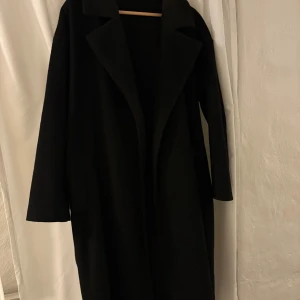 Svart kappa med bälte och krage - Säljer en svart kappa med klassisk krage och bälte i midjan. Jackan har lång ärm och rak passform, perfekt för dig som gillar en stilren look. Materialet är mjukt och kappan har en snygg oversized vibe.
