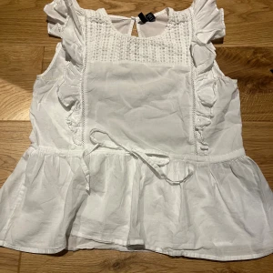 Vit blus med volanger från Vero Moda - Supersöt vit blus från Vero Moda i storlek S. Blusen har volanger längs axlarna, smock upptill, knytband i midjan och en liten knapp i nacken. Tillverkad i mjuk bomull, ärmlös och med en lätt peplum-effekt nedtill. Perfekt för sommarens alla tillfällen!