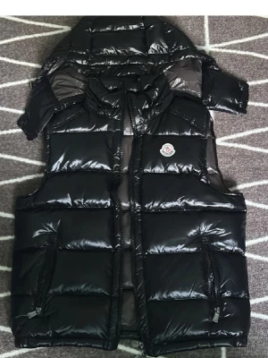 Moncler väst  - Säljer en svart, glansig dunväst från Moncler med huva och dragkedja framtill. Västen har två fickor med dragkedja och klassisk Moncler-logga på bröstet. Insidan har ett coolt serietidningsmönster och Moncler-etikett. Perfekt för lager-på-lager under kyliga dagar.