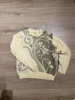 Gul stickad tröja med blommönster - Unik gul stickad tröja med ett stort blommigt mönster i beige och vitt på framsidan. Tröjan har rund hals, ribbade muddar och är mjuk och skön. Perfekt för dig som vill ha något som sticker ut i garderoben.