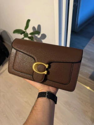 Brun kuvertväska från Coach - Snygg brun kuvertväska från Coach i skinn med guldfärgat spänne framtill. Stilren design med struktur och klassisk form, perfekt för att bära det viktigaste. Väskan har ett elegant och tidlöst uttryck med diskreta sömmar och praktiskt innerfack.