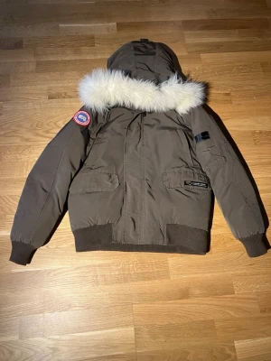 Brun dunjacka från Canada Goose - Snygg brun dunjacka från Canada Goose med stor huva och vit fuskpäls. Jackan har ribbade muddar, två stora fickor framtill och klassisk patch på ärmen. Perfekt för kalla vinterdagar och riktigt varm.