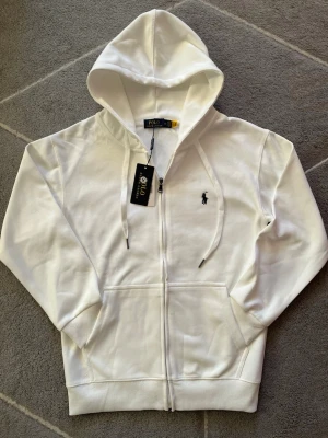 Vit hoodie från Polo Ralph Lauren - Snygg vit hoodie från Polo Ralph Lauren med dragkedja framtill och klassisk huva. Broderad svart logga på bröstet och två fickor framtill. Tillverkad i mjuk bomull för en skön känsla. Perfekt för en avslappnad och stilren look.