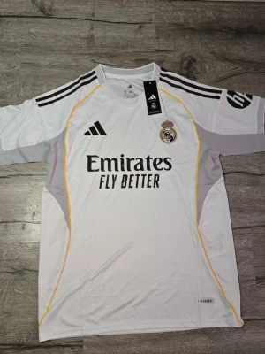 Real Madrid fotbollströja - Säljer en officiell Real Madrid matchtröja från adidas i storlek M. Tröjan är vit med svarta och gula detaljer, Real Madrid-logga på bröstet och klassiska adidas-ränder på axlarna. Materialet är lätt polyester och modellen är slim fit. Perfekt för fotbollsfans!