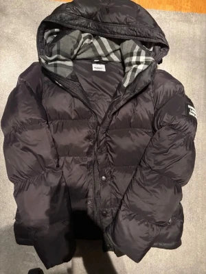 Svart pufferjacka från Burberry - Snygg svart pufferjacka från Burberry med klassiskt rutigt foder i huvan. Jackan har dragkedja och knappar framtill samt en logga på ärmen. Perfekt för kalla dagar och riktigt trendig look.