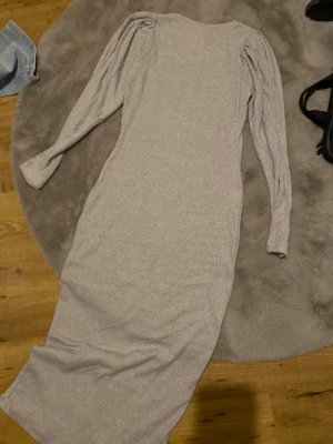 Ljusgrå ribbad långklänning - Långklänning i ljusgrått ribbat material med lång ärm och puffdetaljer vid axlarna. Klänningen har en kroppsnära passform och rund halsringning. Inga defekter.