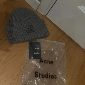 Acne Studios mössa - Säljer en grå ribbstickad mössa från Acne Studios med den klassiska Face-loggan framtill. Mössan är i tjockt, mjukt material och har en bred uppvikt kant. Perfekt för dig som vill ha en stilren och trendig accessoar till hösten och vintern.