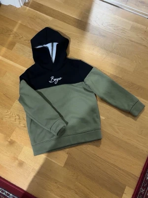 Grön och svart hoodie  - Snygg hoodie i olivgrönt och svart med vit broderad text 'Boys' på bröstet. Hoodien har en klassisk huva och ribbade muddar vid ärmar och nederkant. Tillverkad i mjukt sweatshirtmaterial, perfekt för en avslappnad streetwear-look. Storlek: 8 år