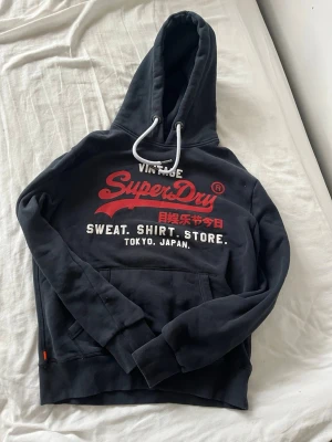 Svart Superdry hoodie med tryck - Snygg svart hoodie från Superdry med stort rött och vitt tryck på bröstet och japanska tecken. Klassisk känguruficka framtill, vita dragsnören i huvan och ribbade muddar. Perfekt för dig som gillar streetwear och vill ha en bekväm och stilren look.