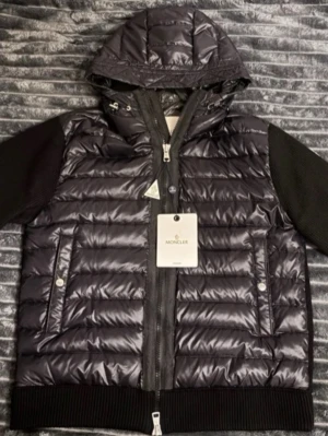 Svart Cardigan med huva från Moncler - Snygg svart Cardigan från Moncler med glansig finish, huva och ribbade stickade ärmar. Jackan har dragkedja framtill, två fickor med tryckknappar och ikonisk Moncler-logga på ärmen. Perfekt för kalla dagar och riktigt stilren look.