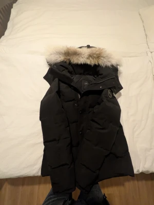 Svart dunjacka med pälskant från Canada Goose - Säljer en svart dunjacka från Canada Goose med stor huva och snygg pälskant. Jackan har klassisk pufferdesign, knappar och dragkedja framtill samt logotyp på ärmen. Perfekt för kalla vinterdagar och riktigt varm.