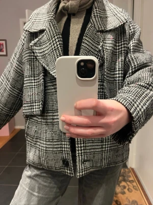  Kort kappa  - Snygg rutig kappa i svart och vitt med bred krage och knappar framtill. Jackan har en loose passform och är långärmad, perfekt för höst och vinter. Materialet känns som ullblandning och mönstret är klassiskt rutigt.