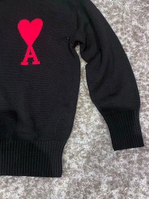 Svart stickad tröja från Ami Paris - Svart stickad tröja från Ami Paris med ikoniskt rött hjärta och bokstaven A broderat på bröstet. Tröjan har rund hals, långa ärmar och breda ribbade muddar vid ärmslut och nederkant. Perfekt för dig som gillar stilrena och trendiga plagg. Har aldrig använt den då jag inte gillar stickade tröjor så mycket, men den var riktigt skön.