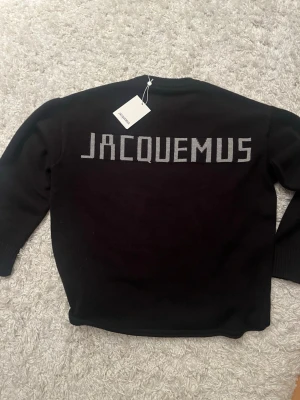 Svart stickad tröja från Jacquemus - Svart stickad tröja från Jacquemus med rund halsringning och ribbade muddar. På ryggen finns en stor Jacquemus-logga i grått. Tröjan har en klassisk, rak passform och är tillverkad i ett mjukt material som känns lyxigt. På andra bilden ser den vinröd ut men detta är endast pga att bilden är tagen med blixt, den har inga missfärgningar.