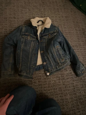 Blå jeansjacka med teddyfoder - Säljer en klassisk blå jeansjacka med mysigt vitt teddyfoder på insidan och kragen. Jackan har knappar framtill, två bröstfickor med lock och två sidofickor. Perfekt för dig som vill ha en tidlös och trendig look.