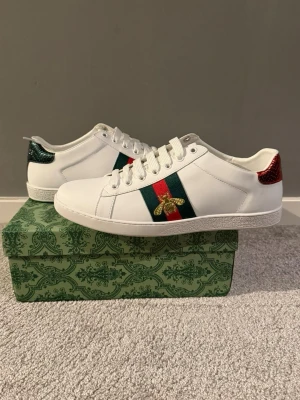 Gucci ace - Säljer ett par vita sneakers från Gucci med klassiska gröna och röda ränder på sidan och en guldbroderad bi. Baksidan har metallicröd och metallicgrön hälflik i ormmönstrat skinn. Skorna har snörning och platt sula.