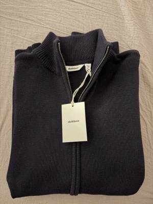 Mörkblå stickad ziptröja Dobber - Snygg mörkblå stickad ziptröja från Dobber med hög krage och ribbade detaljer. Tröjan har dragkedja framtill och är perfekt för lager på lager. Klassisk och stilren design som passar till det mesta.
