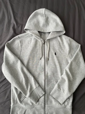 Polo Ralph Lauren zip - Snygg ljusgrå hoodie med dragkedja från Polo Ralph Lauren. Klassisk design med huva, dragsnören och två fickor framtill. Ikoniska svarta Polo-loggan broderad på bröstet. Tillverkad i mjukt bomullsmaterial för maximal komfort.