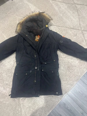Svart parkas från Parajumpers - Säljer en svart parkas från Parajumpers med avtagbar fuskpäls på huvan och flera praktiska fickor. Jackan har långa ärmar, dragkedja och knappar framtill samt patchdetalj på ena ärmen. Perfekt för kalla dagar.