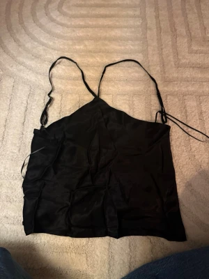 Svart satinlinnetopp med tunna band - Snygg svart linnetopp i satin med tunna axelband och enkel, stilren design. Perfekt till fest. Säljer pga att den inte används .