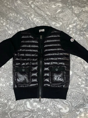 Svart pufferjacka från Moncler - Säljer en svart pufferjacka från Moncler med quiltad framsida och två stora fickor med knappstängning. Jackan har stickade ärmar och rygg, rund halsringning och dragkedja framtill. Klassisk Moncler-logga på ena ärmen. Perfekt för kyliga dagar. OBS! INTE ÄKTA 