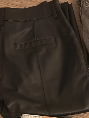 Svarta byxor i skinnimitation - Säljer ett par svarta byxor i skinnimitation med raka ben och medel hög midja. Byxorna har bälteshällor och en söm till bakficka. 