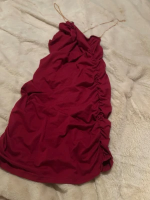 Röd kort klänning med tunna band - Trendig röd kort klänning med tunna axelband i kedja och snygga rynkade detaljer längs sidan. Klänningen har en omlottliknande överdel och är perfekt för dig som vill sticka ut. Materialet känns mjukt och följsamt.