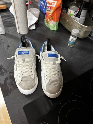 Philippe Model vita sneakers blå detalj - Säljer ett par vita Philippe Model sneakers med blå häl och blå logga på plösen. Skorna har klassisk snörning, rund tå och platt sula. Materialmix av skinn och mocka ger en schysst kontrast. Perfekta för dig som gillar stilrena men ändå unika sneakers. Dom har en lagning på sig