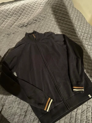 Svart stickad kofta med dragkedja - Säljer en svart stickad kofta från NN07 med hel dragkedja framtill. Tröjan har ribbade muddar och krage samt snygga detaljer i vitt och orange på ärmsluten. Perfekt till höst och vinter, enkel att matcha med det mesta.