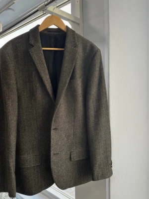 Grå kavaj från River Island, storlek 42 - Snygg grå kavaj från River Island i klassisk modell med två knappar och fickor fram. Materialet är ullmix med diskret fiskbensmönster som ger en stilren look. Perfekt till skjorta eller t-shirt för en clean outfit. Grå/brun färg.