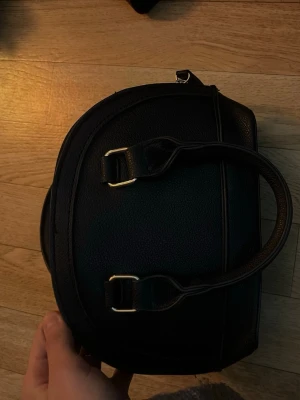 Svart handväska i skinnimitation - Säljer en stilren svart handväska i skinnimitation med rundad form och dubbla handtag. Väskan har guldfärgade metalldetaljer och dragkedja upptill. Perfekt storlek för det viktigaste och enkel att matcha till olika outfits.