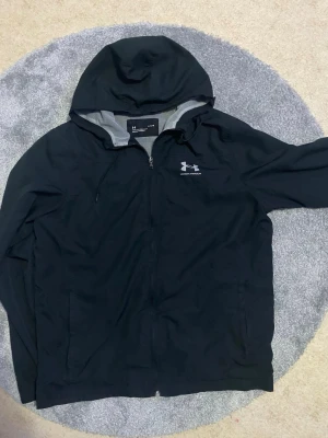Svart vindjacka från Under Armour - Säljer en svart vindjacka från Under Armour med huva och meshfoder. Jackan har dragkedja framtill, två sidofickor och en snygg logga på bröstet. Passformen är loose och materialet är lätt och andas, perfekt för blåsiga dagar.