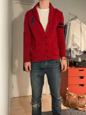 Röd kofta från Ralph Lauren - Säljer en röd kofta från Ralph Lauren med big pony  logga på bröstet och svarta ränder på ena ärmen. Koftan är stickad, har knappar framtill och två fickor. 