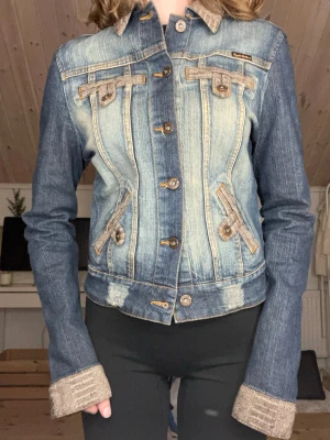 Jeansjacka med snörning från Apple Bottoms - Unik jeansjacka från Apple Bottoms med snygga kontrastsömmar, bruna detaljer och cool snörning i ryggen. Jackan har bruna manchesterdetaljer på krage och ärmslut, broderad logga på ryggen och dekorativa knappar framtill. Perfekt för dig som vill sticka ut.