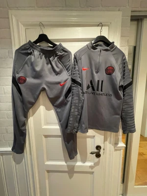 Grå Nike PSG träningsset - Säljer ett grått PSG träningsset från Nike med Dri-FIT material. Setet består av långärmad tröja med halv dragkedja och matchande byxor. Röda Nike-loggor och PSG-emblem på både tröja och byxa, samt svarta detaljer och tryck. Perfekt för fotbollsträning.