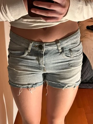 Ljusblå denimshorts med fransar - Säljer ett par ljusblå denimshorts med råa fransar i bensluten och slitna detaljer. Klassisk femficksmodell med knapp och dragkedja framtill. Perfekta för varma dagar och enkel att matcha med olika toppar.