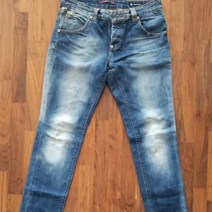 Philipp Plein blå jeans - skull edition - w33 - Snygga blå jeans från Philipp Plein med coola metalldetaljer och döskalleknappar. Jeansen har en faded tvätt, raka ben och klassisk femficksdesign. Orange kontrastsömmar och exklusiva emblem ger en unik look. Perfekt för dig som gillar streetstyle och vill sticka ut.