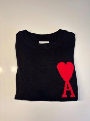 Svart stickad tröja med rött hjärta - Svart stickad tröja från AMI Paris med ett stort rött hjärta och bokstaven A på bröstet. Tröjan har rund halsringning och är tillverkad i ett mjukt, tjockt material som passar perfekt för kyligare dagar.