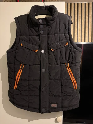 Svart dunväst från Superdry XXL - Svart dunväst från Superdry med orange dragkedjor och detaljer. Västen har två bröstfickor med lock och två snedställda fickor med dragkedja. Insidan är fodrad med mjuk, vit teddy. Perfekt för lager på lager och streetwear.