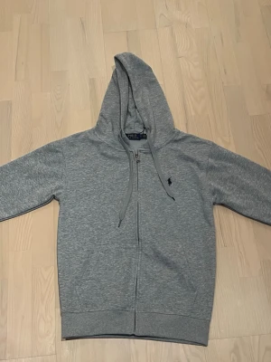 Grå zip hoodie från Polo Ralph Lauren - Snygg grå hoodie från Polo Ralph Lauren och den ser ut att vara mörkgrå men det är den inte utan det är bara ljuset i rummet. Den har dragkedja framtill och klassisk huva med dragsnören. Diskret logga broderad på bröstet. Tillverkad i mjuk bomullsblandning och har ribbade muddar vid ärmar och nederkant. Perfekt för en chill och stilren look.