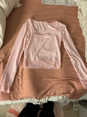 Ljusrosa långärmad topp med spets - Söt ljusrosa långärmad topp med spetsdetaljer vid halsringning, ärmslut och nederkant. Toppen har en fyrkantig ringning och croppad passform. 