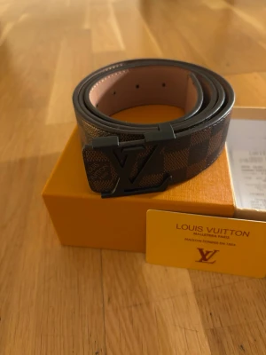 Louis Vuitton bälte brun/svart - Snyggt bälte från Louis Vuitton i klassiskt Damier-mönster i brunt och svart. Bältet har ett stort LV-spänne i svart metall och är gjort i läder med exklusiv känsla. Perfekt accessoar för att lyfta din outfit med en lyxig touch.