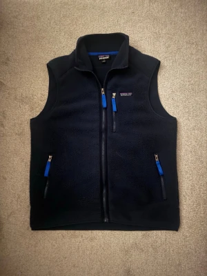 Mörkblå fleeceväst från Patagonia - Mysig mörkblå fleeceväst från Patagonia med hel dragkedja och tre fickor med blå detaljer på dragkedjorna. Klassisk logga på bröstet och hög krage för extra värme. Perfekt lager för kyliga dagar och outdoor-häng. Den sitter som L.