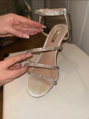 Glittriga klackar med smala remmar - Snygga klackar i silver med flera smala remmar dekorerade med glittriga stenar. Skon har öppen tå, hög klack och justerbart spänne runt ankeln. Perfekta för att lyfta din outfit med extra bling. Klackhöjd 9,5